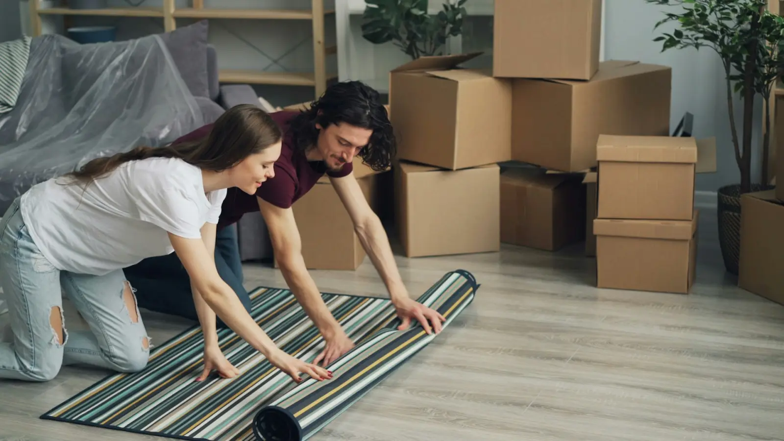 Junges Paar rollt in der neuen Wohnung einen Teppich aus, daneben Umzugskartons