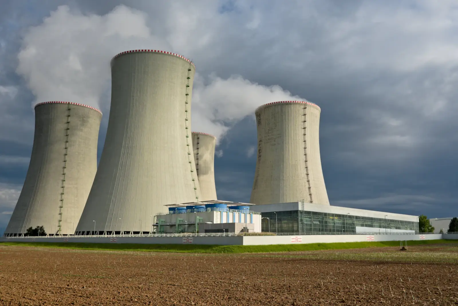 Vier Kühltürme eines Kernkraftwerks mit aufsteigendem Wasserdampf, davor ein langgestrecktes Betriebsgebäude, im Vordergrund ein gepflügtes Feld unter bewölktem Himmel.